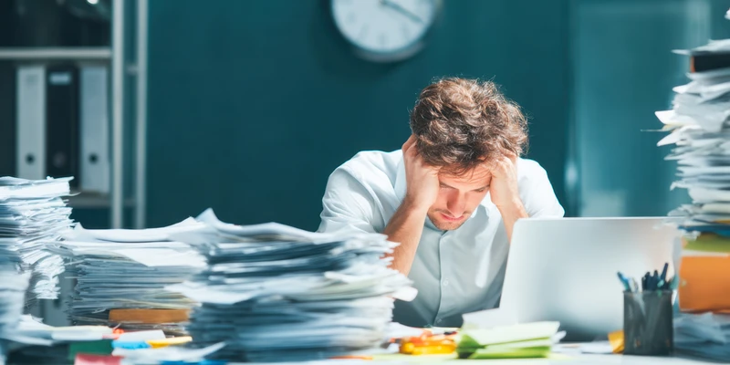 Overwhelmed man sitting at a desk surrounded by piles of paperwork, holding his head while working on a laptop, representing stress, burnout, and mental pressure commonly felt during Blue Monday.