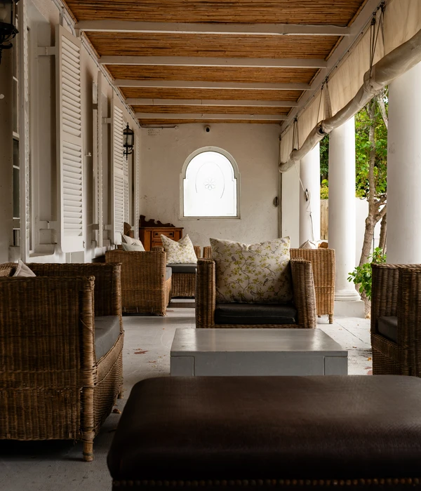 A long covered terrace with wicker armchairs, floral pillows, white pillars, and a bamboo-style ceiling.