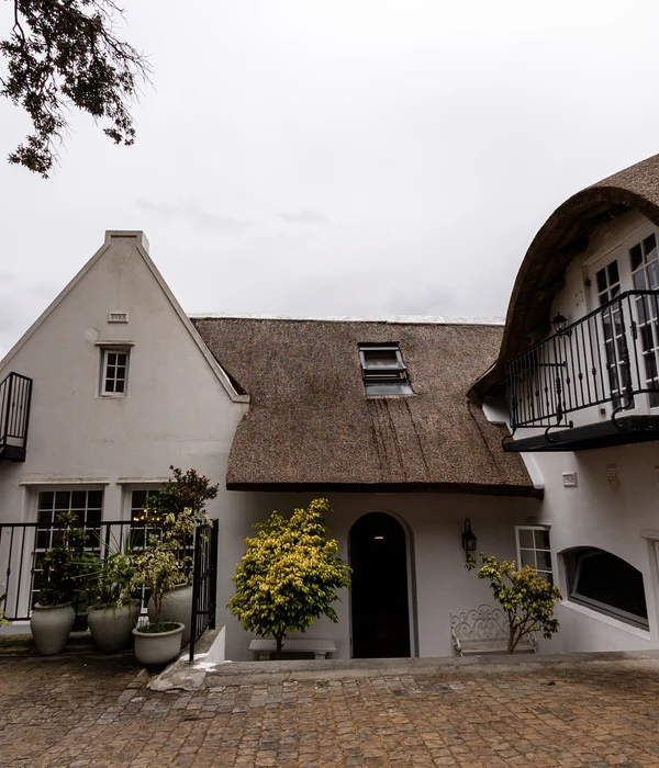 Courtyard of a white manor with thatched roofs, cobblestone ground, potted plants, and a small arched doorway.