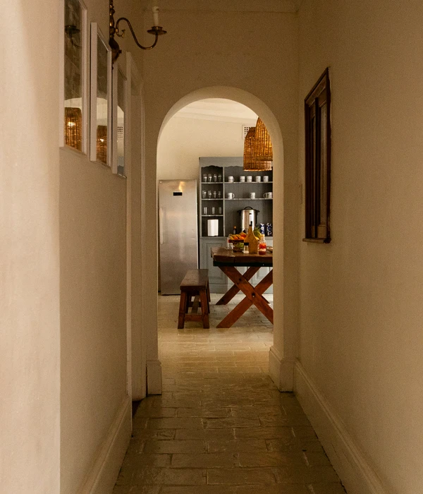 View looking down a hallway through an arched doorway into a rustic kitchen with a wooden table, bench, and stone floors.