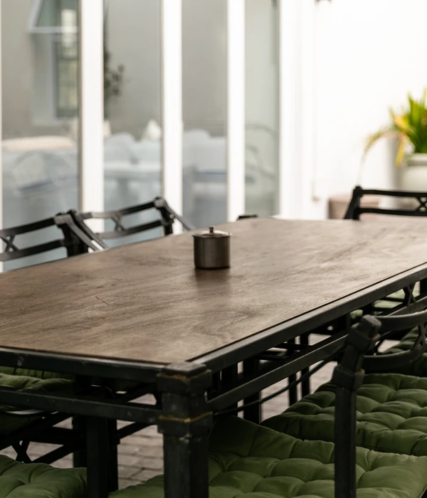 An outdoor wooden dining table with black metal chairs and green cushions, set in a bright, tranquil courtyard area.