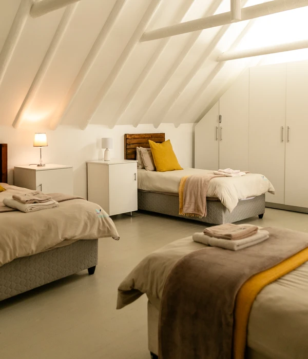 A shared attic bedroom with three neatly made beds, yellow accents, and white wardrobes under a beamed vaulted ceiling.