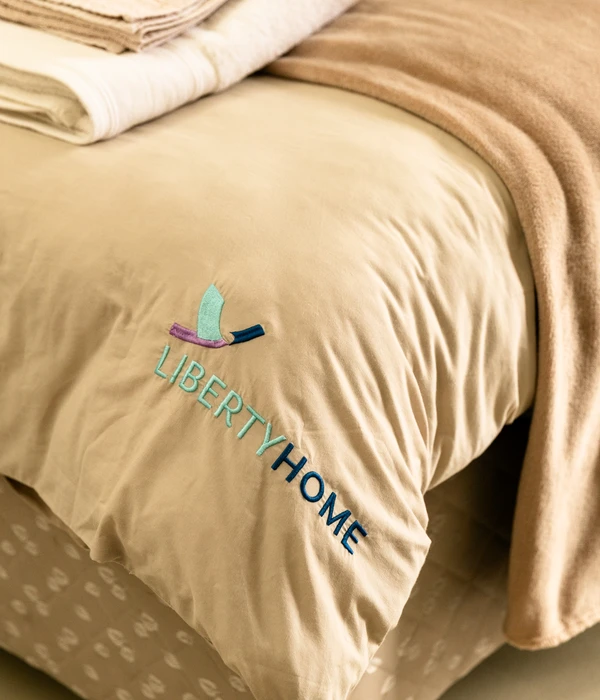 Close-up of a neatly made bed with beige linens featuring an embroidered "LIBERTY HOME" logo and folded towels.