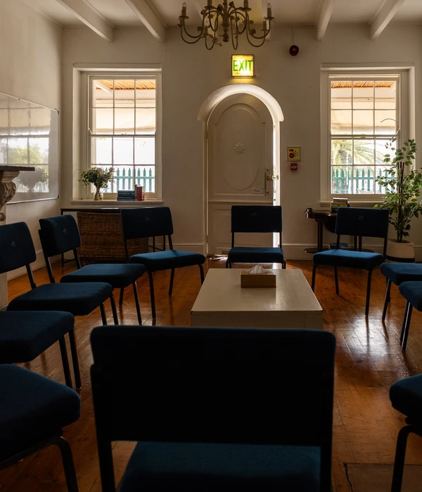 A sunlit group therapy room with blue chairs arranged in a circle around a low table with tissues on a wooden floor.