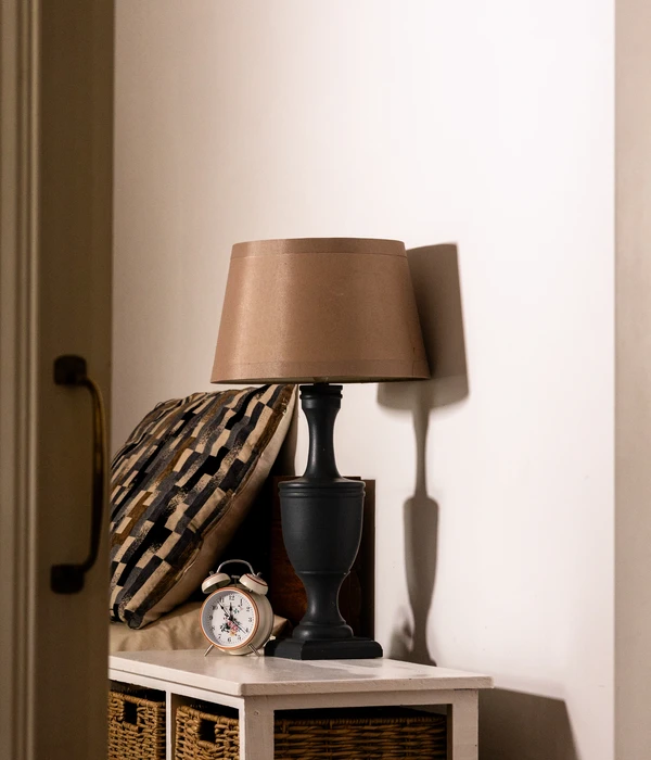 A warm bedside lamp glows on a white nightstand next to a vintage alarm clock and a patterned pillow.