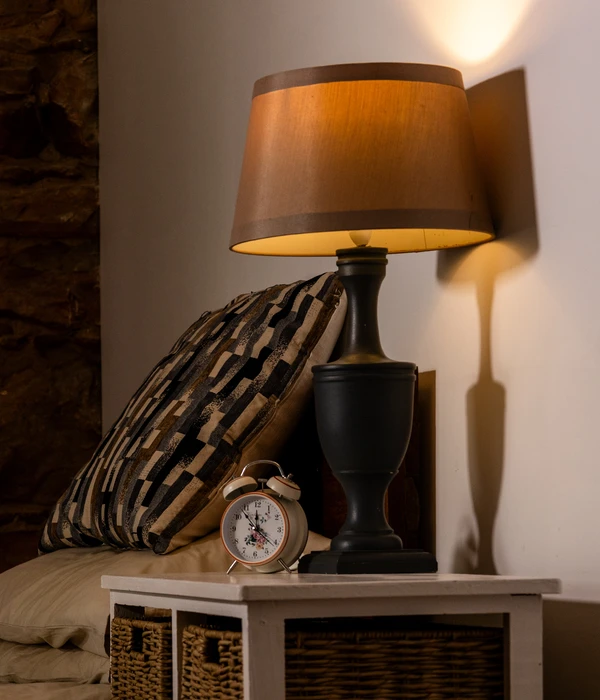 A warm bedside lamp glows on a white nightstand next to a vintage alarm clock and a patterned pillow.