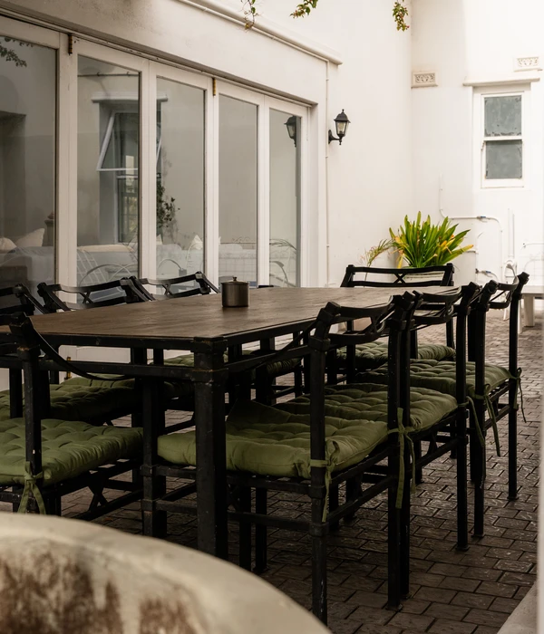 An outdoor wooden dining table with black metal chairs and green cushions, set in a bright, tranquil courtyard area.