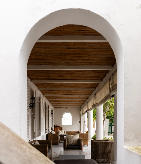 A long covered terrace with wicker armchairs, floral pillows, white pillars, and a bamboo-style ceiling.