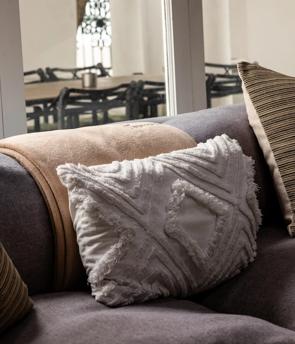 Close-up of a comfortable grey sofa with a textured white pillow and a beige blanket, overlooking a bright dining area.
