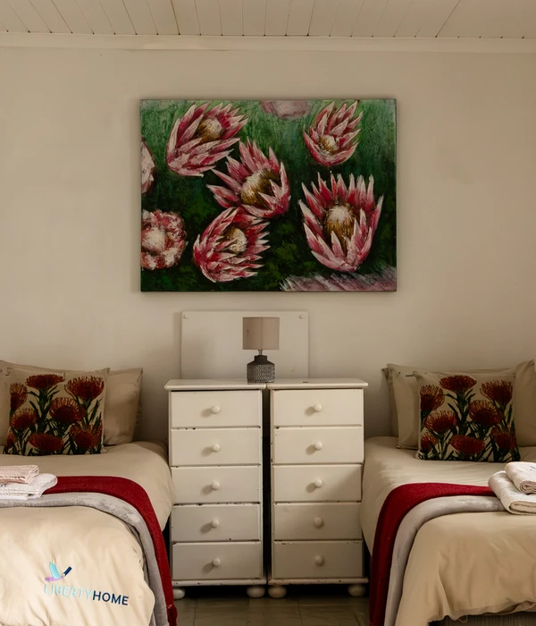 Two twin beds with red throws flanking a white dresser, with a large protea flower painting hanging on the wall above.